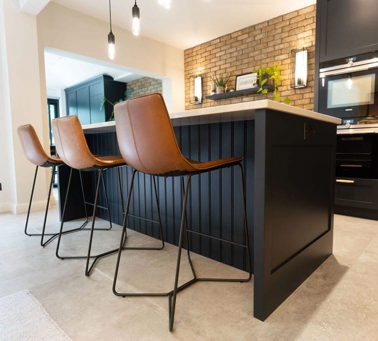 Close-up view of a kitchen island with a white countertop and brown leather bar stools, set against dark blue cabinetry and exposed brick walls.