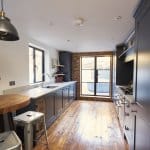 Narrow kitchen with dark blue shaker cabinets, white countertops, and wooden flooring, featuring a round wooden table and metal stools, with a window and a glass door at the far end.