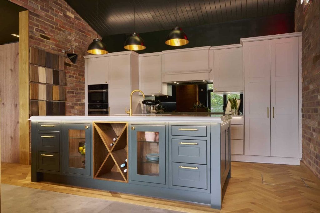 Kitchen in showroom with a blue island featuring glass and wooden storage sections, white cabinetry, and exposed brick walls, illuminated by pendant lights.