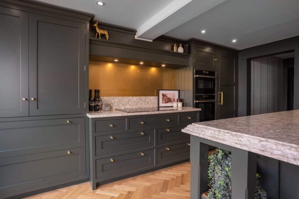 A kitchen with dark grey shaker cabinetry, brass hardware, and a marble countertop, featuring a gold backsplash and wooden herringbone flooring.